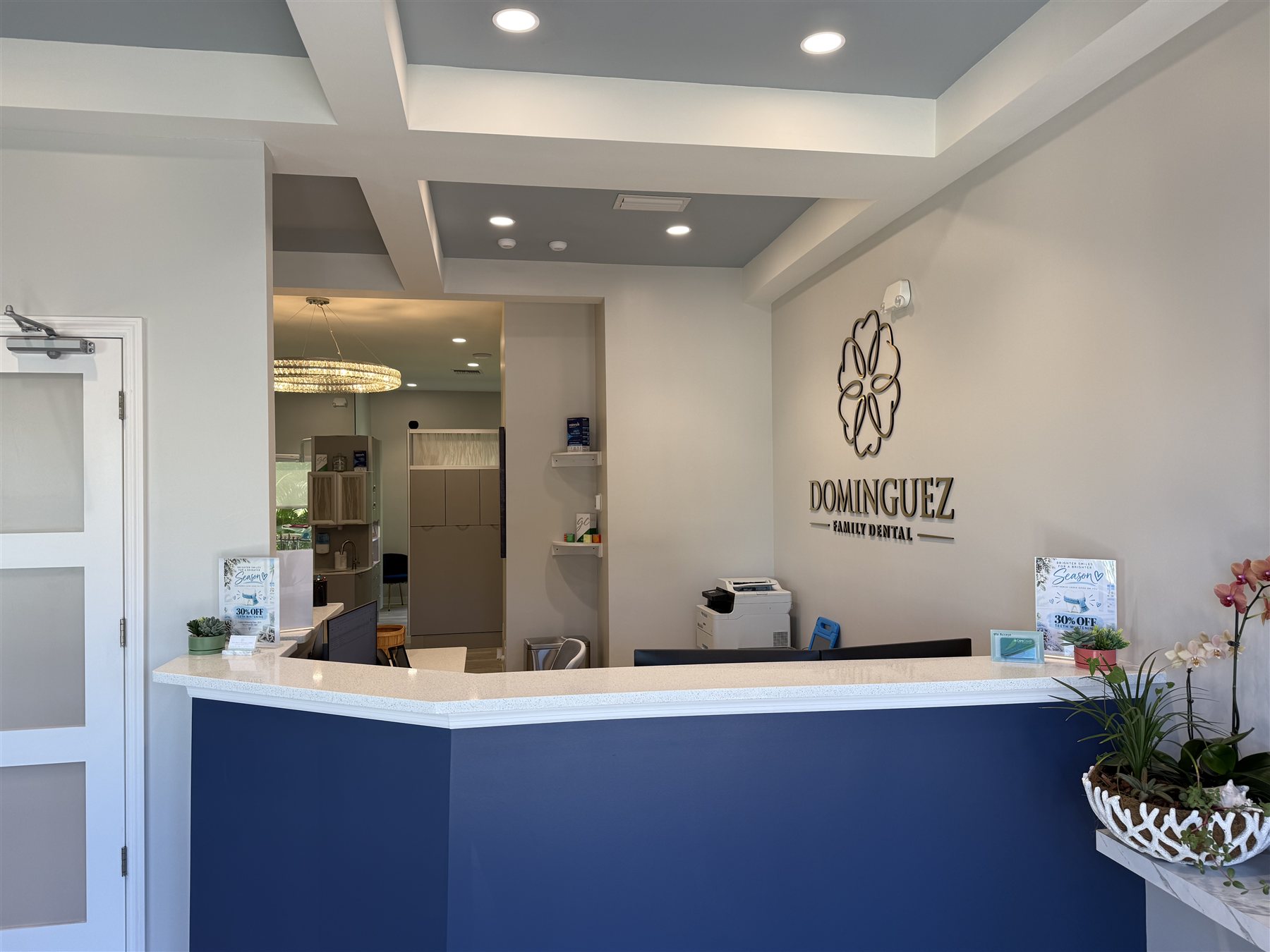 The front desk with a blue front, white countertop, orchids, and the Dominguez Family Dental logo on the wall.